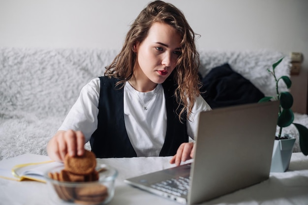 Mujer joven viendo series o podcasts mirando la pantalla con interés sosteniendo galletas en la mano