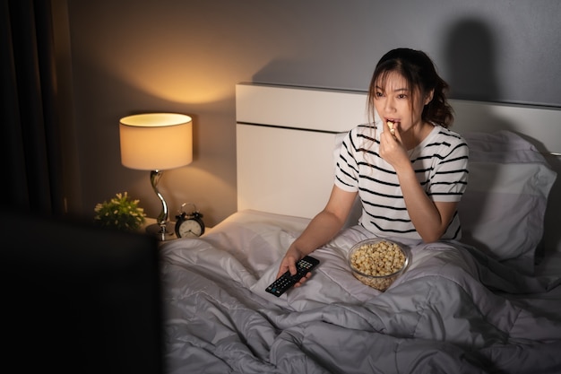 Mujer joven viendo la televisión y comiendo palomitas de maíz en una cama por la noche