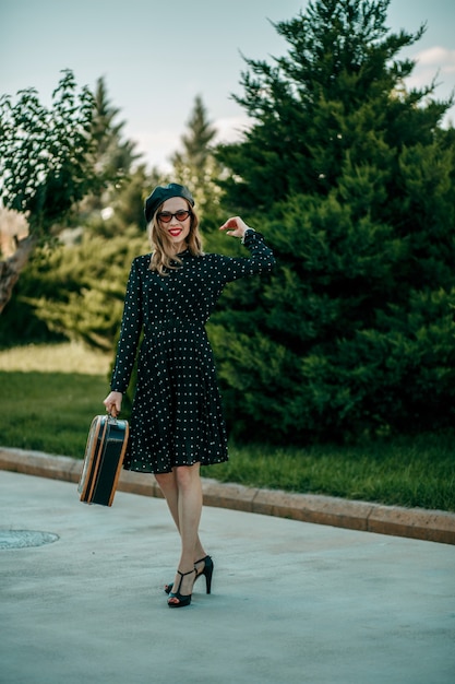 Mujer joven en vintage vestido de lunares negros con maleta retro en la mano posando afuera