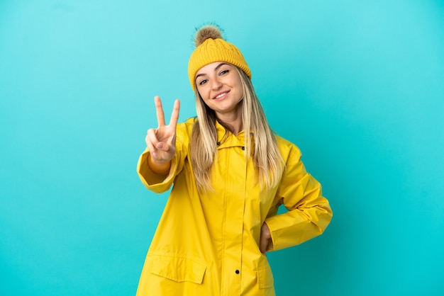 Mujer joven vistiendo un abrigo impermeable sobre fondo azul aislado sonriendo y mostrando el signo de la victoria