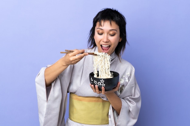 Mujer joven vistiendo kimono sobre pared azul aislado sosteniendo un tazón de fideos con palillos