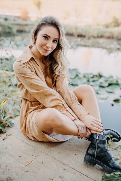 Mujer joven vistiendo un maxi vestido de gasa sentado en un muelle de madera junto a un estanque mirando a la cámara sonriendo