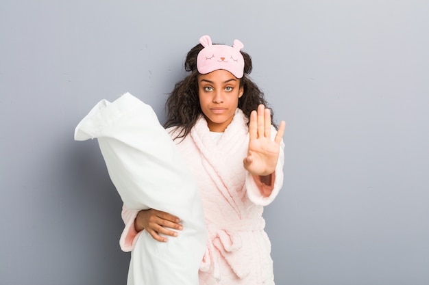 Mujer joven vistiendo pijamas y una máscara para dormir sosteniendo una almohada de pie con la mano extendida que muestra la señal de stop