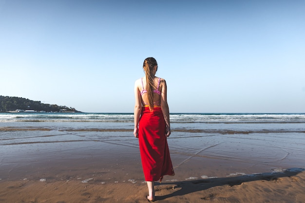 Mujer joven vistiendo una toalla roja y un bikini rosa caminando al borde del agua en la playa