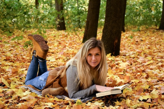 Mujer joven yace en el parque sobre hojas amarillas y lee un libro