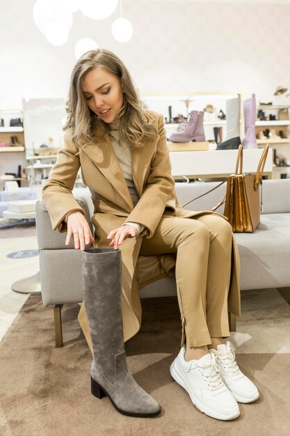 Una mujer joven en una zapatería elige botas Una hermosa rubia con un elegante abrigo beige se sienta en un puf y se prueba los zapatos de otoño Cambio estacional de colección Moda y estilo Vertical