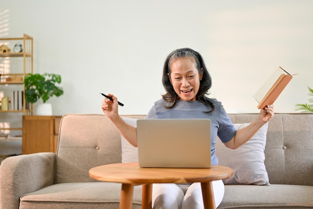 Mujer jubilada asiática emocionada y feliz usando su computadora portátil en la sala de estar ganando la lotería