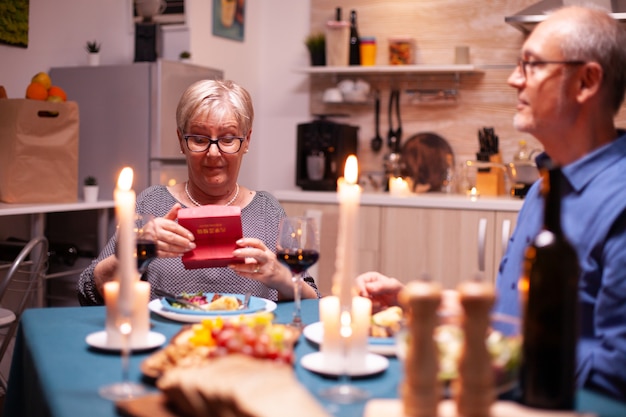 Mujer jubilada con caja de regalo del marido durante la cena de aniversario de la relación. Feliz alegre pareja de ancianos cenando juntos en casa, disfrutando de la comida, celebrando su matrimonio, sorpresa holid