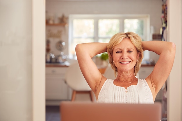 Mujer jubilada en casa en la cocina feliz con sentido de logro al final del día trabajando en la computadora portátil