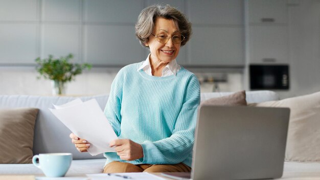 Mujer jubilada feliz sentada usando una computadora y sosteniendo papeles leyendo correos electrónicos y comprobando facturas