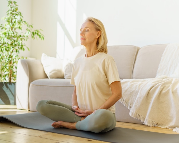 Mujer jubilada meditando y practicando yoga mientras está sentado en posición de loto en el piso en casa