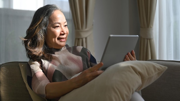 Mujer jubilada sentada en un cómodo sofá y revisando las noticias por correo electrónico en línea en la tableta Concepto de tecnología para personas mayores