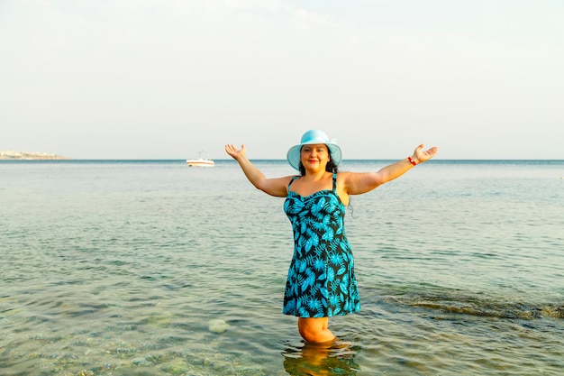 Una mujer judía feliz con un vestido azul y un sombrero se encuentra en el mar hasta las rodillas en el agua. Foto horizontal