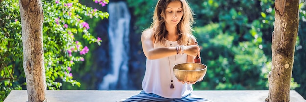 Mujer jugando en un cuenco tibetano mientras está sentado en una alfombra de yoga contra una cascada vintage tonificada