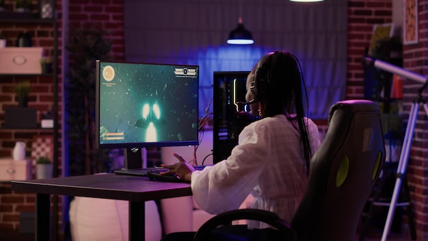 Mujer jugando simulación de disparos espaciales multijugador disfrutando del tiempo libre usando la configuración de juegos de pc en la sala de estar de casa. Chica jugadora afroamericana hablando con auriculares mientras transmite un juego de acción en línea.