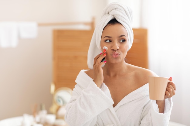 Mujer juguetona aplicando crema hidratante en la cara con café en el baño