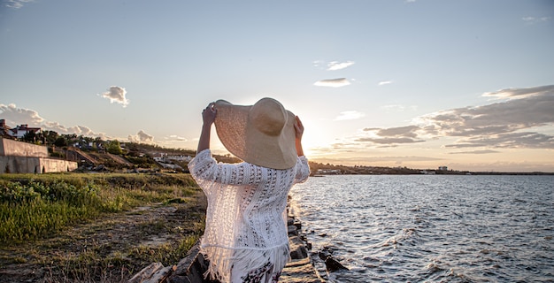 Foto mujer junto al mar al atardecer vista posterior. vestido con falda larga y sombrero.