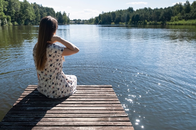 Foto mujer en el lago. mujer joven con vestido blanco disfrutando de la naturaleza y descansando sentado en un muelle de madera en el lago, vista posterior