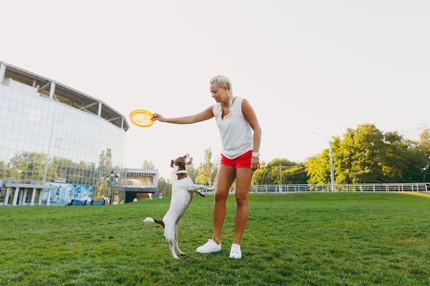 Mujer lanzando un disco volador naranja a un pequeño perro gracioso, que lo atrapa en la hierba verde. Pequeña mascota Jack Russel Terrier jugando al aire libre en el parque. Perro y dueño al aire libre.