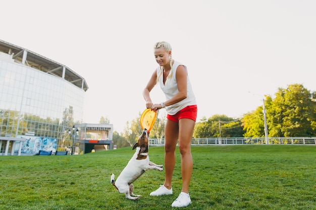 Mujer lanzando un disco volador naranja a un pequeño perro gracioso, que lo atrapa en la hierba verde. Pequeña mascota Jack Russel Terrier jugando al aire libre en el parque. Perro y dueño al aire libre.