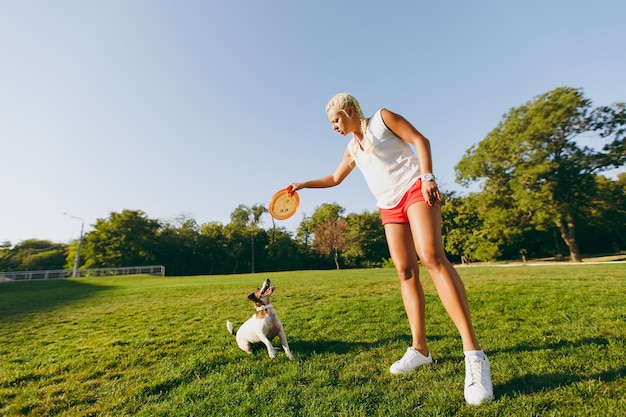 Mujer lanzando un disco volador naranja a un pequeño perro gracioso, que lo atrapa en la hierba verde. Pequeña mascota Jack Russel Terrier jugando al aire libre en el parque. Perro y dueño al aire libre.