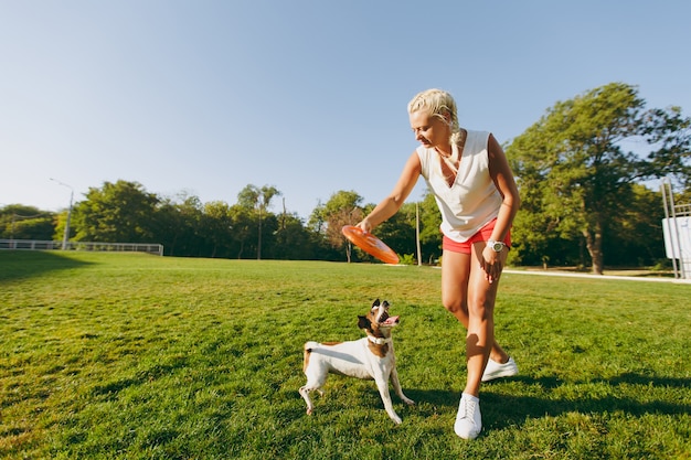 Mujer lanzando un disco volador naranja a un pequeño perro gracioso, que lo atrapa en la hierba verde. Pequeña mascota Jack Russel Terrier jugando al aire libre en el parque. Perro y dueño al aire libre.