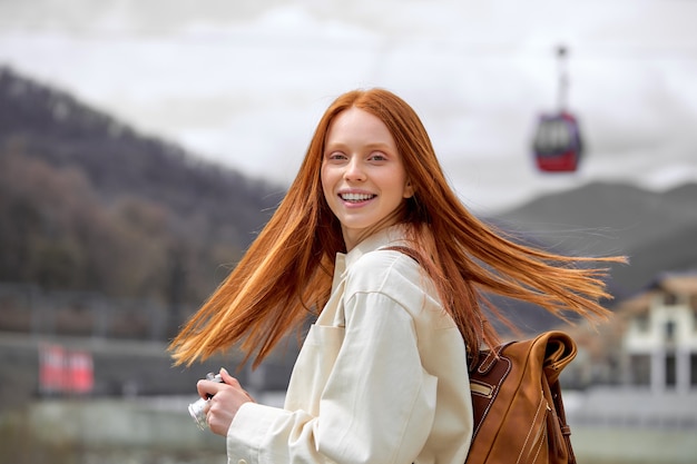 Mujer con largo cabello lacio rojo vistiendo chaqueta beige de pie entre el paisaje de las montañas, mira a la cámara, girando la cabeza hacia atrás. Copie el espacio. Retrato