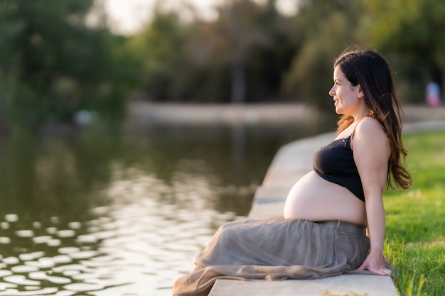 Mujer latina adulta embarazada contemplando el río de un parque mientras está sentada en el borde con una expresión feliz vistiendo ropa cómoda de verano durante la puesta de sol