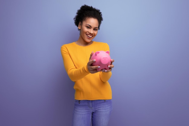 Una mujer latina bastante alegre con cabello afro en un suéter amarillo informal acumuló dinero en un cerdito