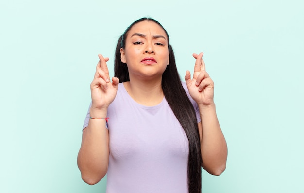 Mujer latina cruzando los dedos con ansiedad y esperando buena suerte con una mirada preocupada