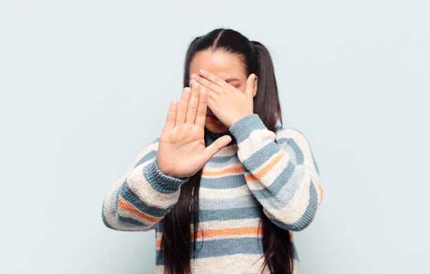 Mujer latina cubriendo la cara con la mano y poniendo la otra mano al frente para detener la cámara, rechazando fotos o imágenes