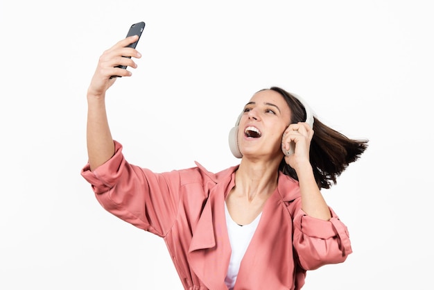 Mujer latina feliz con auriculares bailando y tomando un selfie sobre fondo blanco.