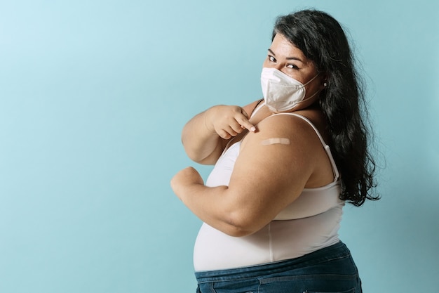 Foto mujer latina de gran tamaño con mascarilla apuntando con su dedo una vacuna de vendaje