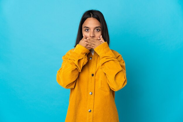 Mujer latina joven aislada en la pared azul que cubre la boca con las manos