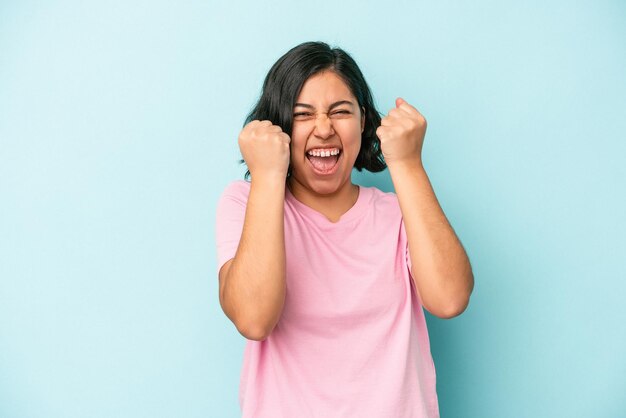 Mujer latina joven aislada sobre fondo azul animando despreocupada y emocionada. Concepto de victoria.