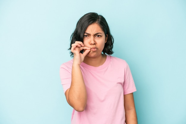 Mujer latina joven aislada sobre fondo azul con los dedos en los labios guardando un secreto.