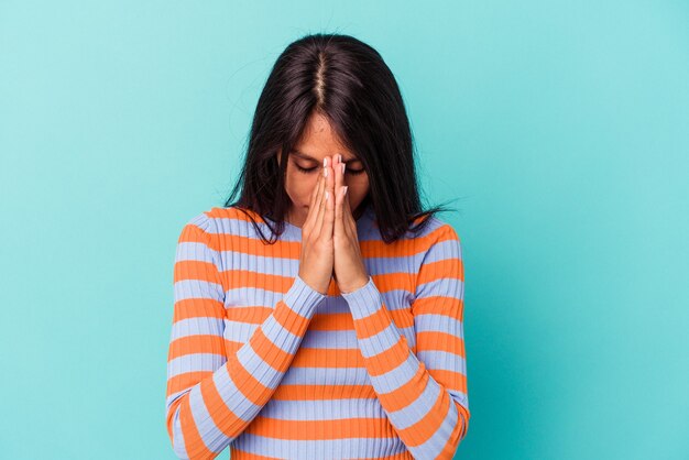 Mujer latina joven aislada sobre fondo azul rezando, mostrando devoción, persona religiosa en busca de inspiración divina.