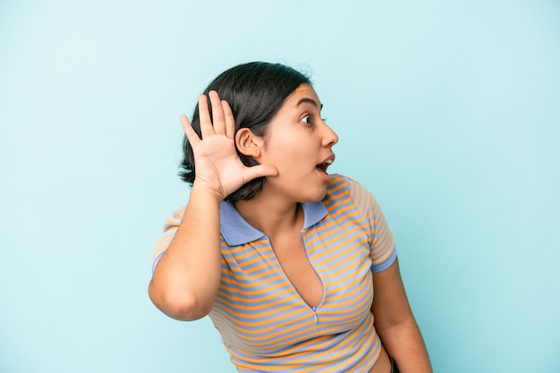 Mujer latina joven aislada sobre fondo azul tratando de escuchar un chisme.