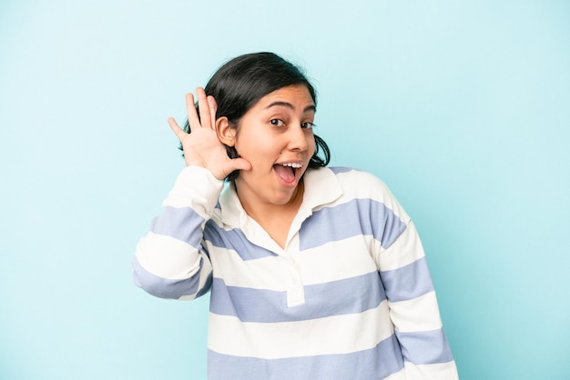 Mujer latina joven aislada sobre fondo azul tratando de escuchar un chisme.