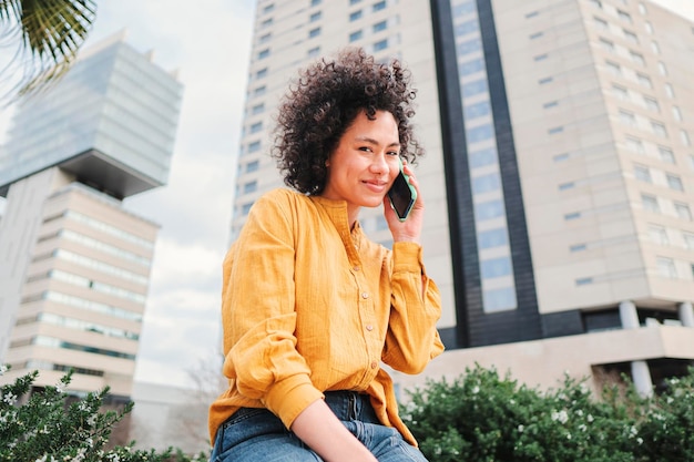 Mujer latina joven con cabello rizado que tiene una llamada por teléfono inteligente Mujer hispana con camisa amarilla hablando por teléfono celular sentado al aire libre Concepto de comunicación