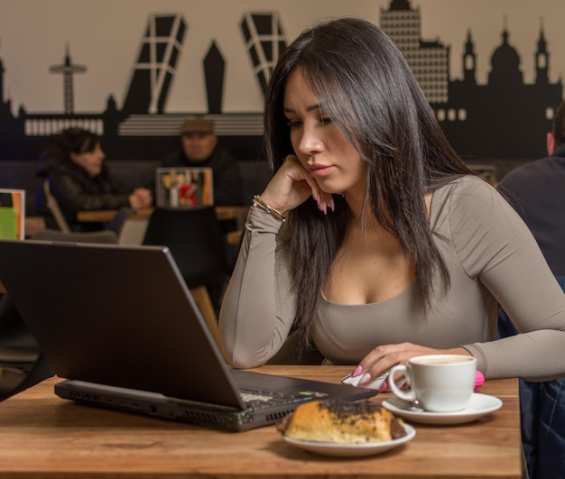 Mujer latina joven en camiseta marrón trabajando o estudiando en una computadora portátil, una computadora, un café, sentada en la mesa.