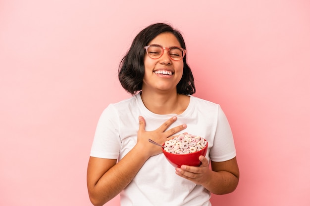 Mujer latina joven con cereales aislados sobre fondo rosa se ríe a carcajadas manteniendo la mano en el pecho.