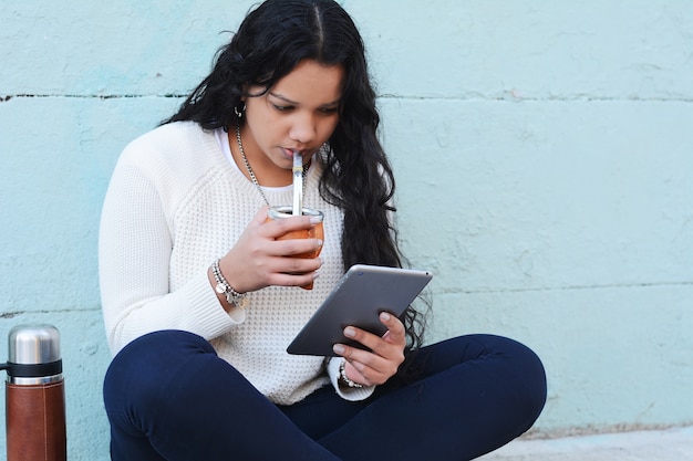 Mujer latina joven que bebe té tradicional de la yerba mate con la tableta