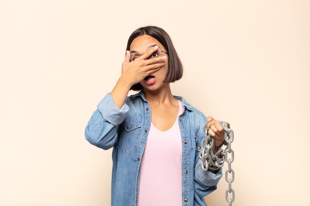 Mujer latina joven que parece sorprendida, asustada o aterrorizada, cubriendo la cara con la mano y mirando entre los dedos