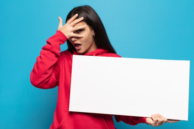 Mujer latina joven que parece sorprendida, asustada o aterrorizada, cubriendo la cara con la mano y mirando entre los dedos