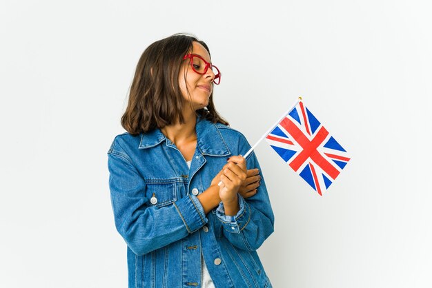 Mujer latina joven que sostiene una bandera inglesa aislada en abrazos de pared blanca, sonriendo despreocupada y feliz.