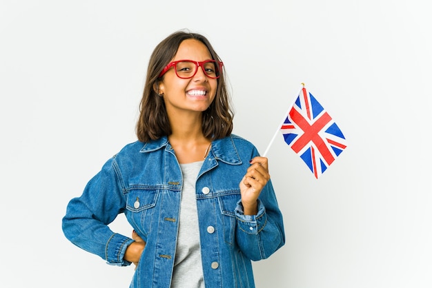 Mujer latina joven que sostiene una bandera inglesa aislada en la pared blanca confiada manteniendo las manos en las caderas.