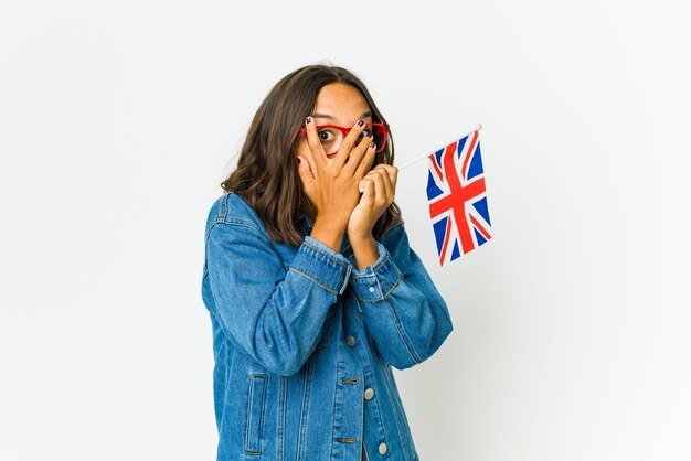 Mujer latina joven que sostiene una bandera inglesa aislada en la pared blanca parpadea entre los dedos asustada y nerviosa.