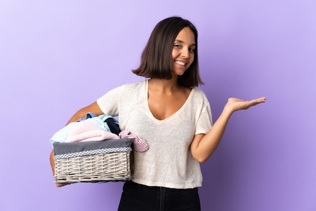 Mujer latina joven que sostiene una cesta de ropa aislada en púrpura que presenta una idea mientras mira sonriente hacia