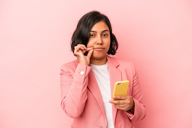 Mujer latina joven que sostiene el teléfono móvil aislado en fondo rosado con los dedos en los labios que guardan un secreto.